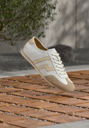 Weißer und beiger Ledersneaker mit Schnürsenkeln, der über einer Oberfläche aus Holzblöcken und Kieseln schwebt, mit verschwommenem Grünbewuchs und Betonhintergrund.