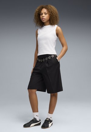 Kobieta o kręconych włosach, ubrana w biały bezrękawnik, czarne szorty do kolan z łańcuchem z gwiazdek na pasku, białe skarpetki i czarne sneakersy marki Puma.
