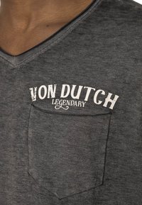 T-shirt gris à col en V en tissu doux, avec une poche frontale. Le texte imprimé "VON DUTCH LEGENDARY" est en blanc avec un effet usé.