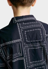 Camisa negra con patrones de pañuelo blanco, con cuello, detalle de botones y un bolsillo parche en la parte posterior, hecha de tela ligera.