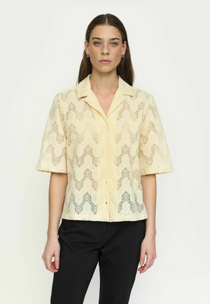 Fiatal nő, hosszú barna hajjal, rövid ujjú, bézs színű csipkés inggombolással és fekete nadrágot visel, egy egyszerű, világos háttér előtt állva.