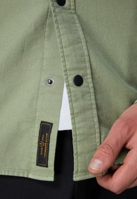 Green textured shirt with black snap buttons, featuring a black label with orange text, partially unbuttoned to reveal a white undershirt.