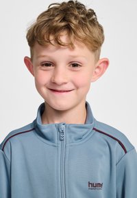 Jasnoniebieska kurtka zapinana na zamek błyskawiczny, z wysokim kołnierzem, z bordo paskiem na ramionach oraz logo na lewej piersi.