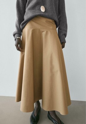 Frau trägt einen lockeren beigen Midirock, grauen Pullover, schwarze Ballerinas und eine ovale Anhängerhalskette, steht vor einem schlichten hellen Hintergrund.