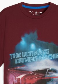 T-shirt bordeaux avec un graphisme d'une voiture de course, entourée de fumée et de lumières bleues, accompagné du texte "LA MACHINE DE CONDUITE ULTIME."
