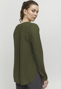 Olive green long-sleeve blouse with a rounded hem, back pleat detail, and a soft, smooth texture; styled with a subtle sheen.