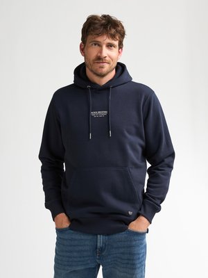 Hombre con las manos en los bolsillos, vistiendo una sudadera con capucha azul marino con el logo de Petrol Industries y vaqueros azules sobre un fondo claro y liso.