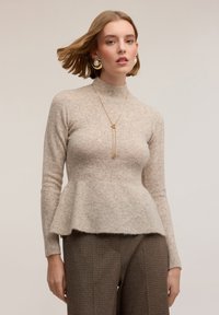 Béžový peplum svetr s vysokým límcem, vyrobený z měkké pletené látky. Kombinován s texturovanými károvanými kalhotami a zlatými šperkovými detaily.