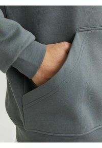 Tejido de sudadera gris con una textura suave, que presenta un bolsillo frontal y puños acanalados. La mano está colocada dentro del bolsillo.