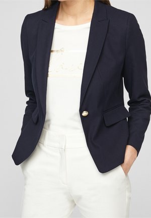 Blazer marine ajusté, avec un bouton doré unique et des revers pointus, porté sur un top blanc avec un imprimé doré "love", associé à un pantalon blanc.