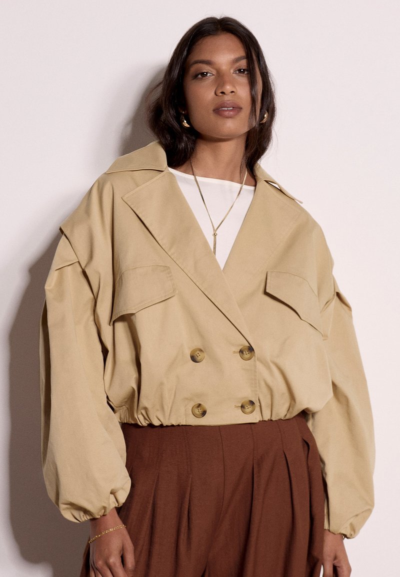 Femme portant une veste courte oversize beige avec de gros boutons et un pantalon plissé marron taille haute, debout devant un fond uni.