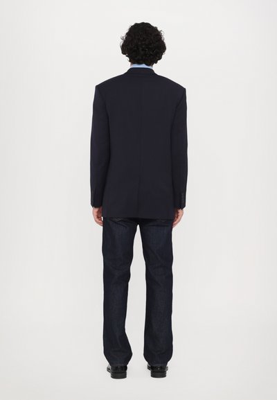 Blazer marine à épaules structurées, associé à un jean en denim foncé. Le blazer présente une fente simple et des poignets boutonnés, créant un look ajusté.