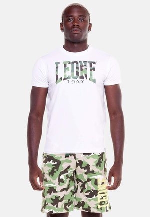 Uomo in piedi che indossa una t-shirt bianca con la scritta "LEONE 1947" e pantaloncini mimetici verdi con la scritta gialla "LEONE" sulla gamba sinistra.