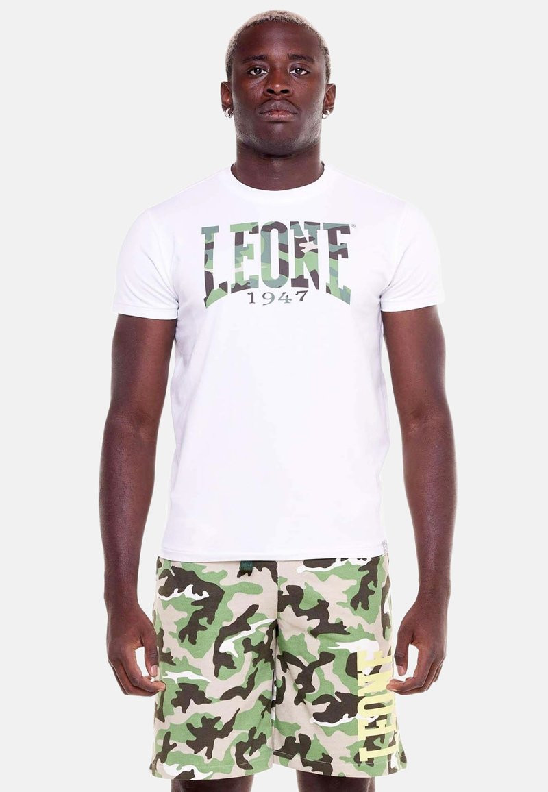 Homme debout portant un t-shirt blanc avec le texte «LEONE 1947» et un short camouflage vert avec le texte jaune «LEONE» sur la jambe gauche.