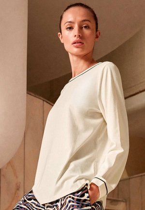 Femme portant un chemisier crème à manches longues avec une bordure noire et un pantalon à motifs, debout avec une main dans la poche contre un fond neutre.
