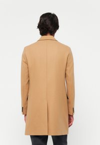 Palton de cămilă cu un design ajustat, guler tăiat, ventral în spate și accente cu nasturi pe mâneci. Textură netedă, asemănătoare lanii.