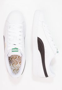 Chaussure avec une tige synthétique blanche, une bande latérale marron, une semelle en caoutchouc texturée et une semelle intérieure beige à motif. Fermeture traditionnelle à lacets.