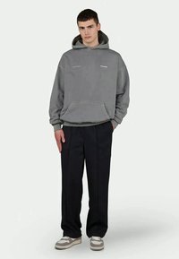 Hanorac gri cu glugă, dotat cu un buzunar frontal și cu branding subtil, purtat împreună cu pantaloni negri largi și adidași bej.