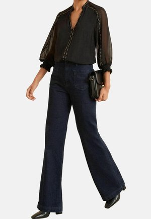 Personne portant un chemisier noir transparent avec un bord clouté, un jean large foncé, des chaussures pointues noires, tenant un sac à main noir dans la main droite.