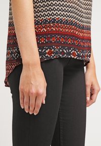 Femme portant un haut à motifs géométriques et rouges, ainsi qu'un pantalon noir, les mains détendues le long du corps, sur un fond uni.