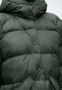 Veste matelassée vert foncé avec fermeture à boutons-pression et col montant, montrant des sections matelassées et des poches avant inclinées.