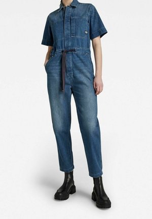 Salopette en denim bleu foncé, à manches courtes, avec poche poitrine, taille ceinturée, jambes fuselées, associée à des bottes noires à semelles épaisses. Texture lisse.