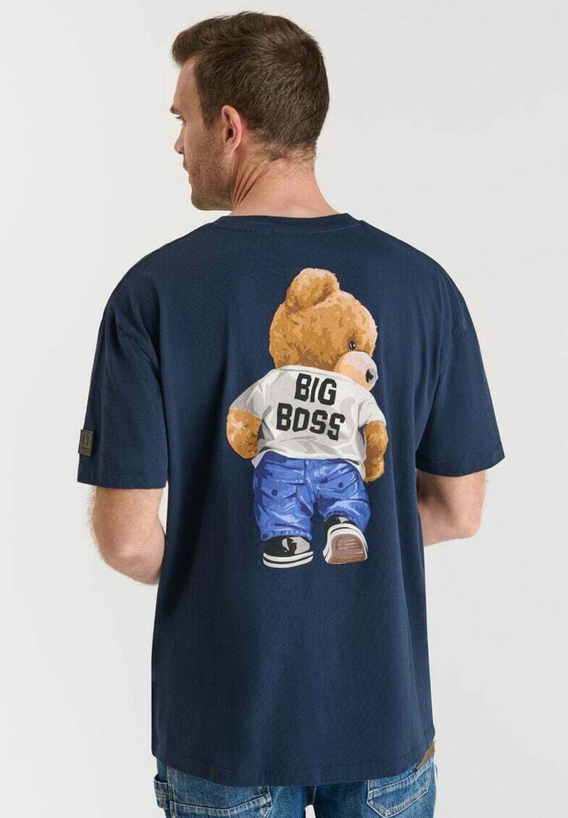 Man in een marineblauwe t-shirt met een afbeelding van een teddybeer in jeans en sneakers, witte shirt met het label "BIG BOSS" op de achterkant.