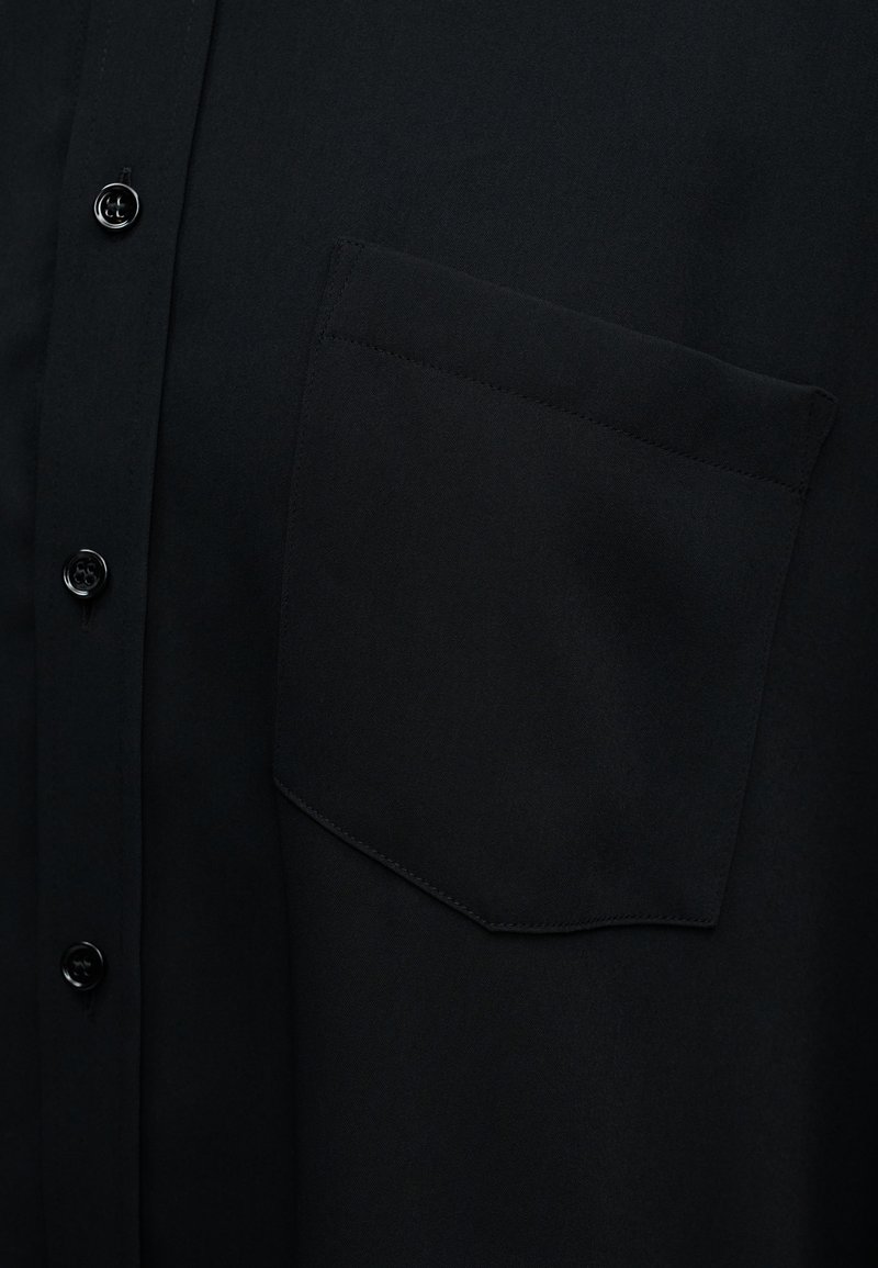 Chemise noire boutonnée avec une texture lisse, dotée d'une poche poitrine et de boutons ronds. Le tissu est léger et uni, sans motifs.