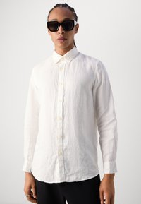 Chemise blanche en lin à manches longues avec un col bouton-down, présentant une finition texturée et un ourlet droit, associée à des lunettes de soleil rectangulaires noires.