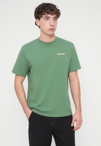Green cotton t-shirt with short sleeves, crew neck, and a small gold Timberland logo on the left chest. Smooth texture, casual fit.