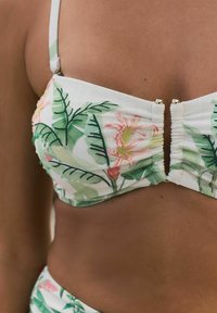 Florallinen bikinitoppi, jossa on vihreitä lehtiä ja vaaleanpunaisia kukkia valkoisella taustalla, röyhelömuotoilu ja kultaiset yksityiskohdat.