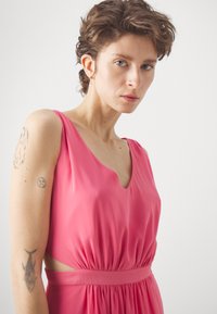 Rožinė beprotė su V formos iškirpte, susirinkusia juosmeniu ir išpjautomis šoninėmis dalimis. Lygus audinys su tekantis drape. Ryški tatuiruotė ant rankos.