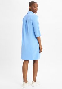 Abito azzurro chiaro con colletto, maniche tre quarti e piega sul retro. Indossato con sneaker bianche. Il materiale appare liscio e casual.