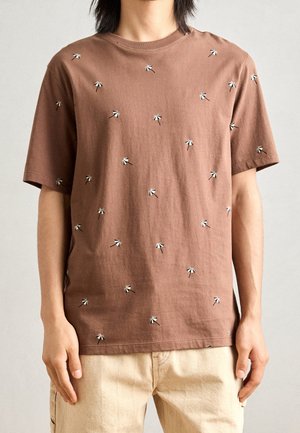 Person iført en brun kortærmet t-shirt med små hvide og sorte paraplymønstre, sammen med beige bukser, stående mod en ensfarvet baggrund.