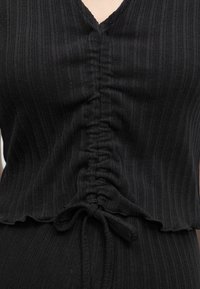 Μαύρο τοπ από ριπ ύφασμα με λαιμόκοψη σε σχήμα V και κεντρικό ρυθμιζόμενο κορδόνι δεμένο με φιόγκο στη μέση.