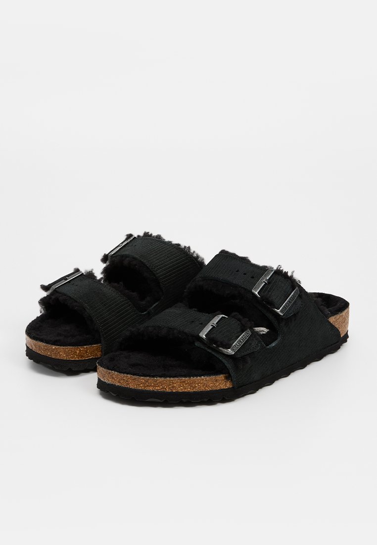 Sandales noires à semelle plate avec doublure en fourrure, deux sangles réglables et boucles en métal. Semelle en liège avec une semelle en caoutchouc texturée pour l'adhérence.