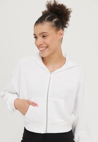 Bijela zip-up hoodie izrađena od mekanog materijala. Ima prednji kenguruh džep, opušteni kroj i kapuljaču s vezicom.