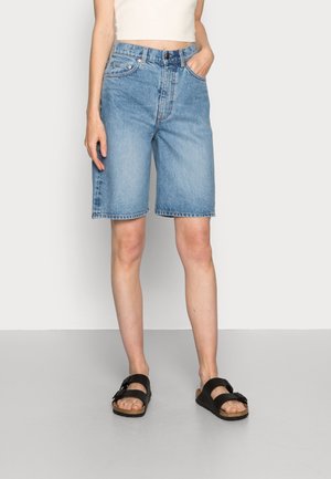 Persona que lleva pantalones cortos de mezclilla azul claro hasta la rodilla, top blanco corto y sandalias negras con dos tiras, de pie sobre un suelo gris frente a una pared blanca.