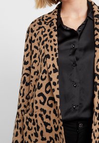 Svetle hnědý cardigan s leopardím potiskem vyrobený z jemné tkaniny, s černými skvrnami, s vykrojeným límcem a předními kapsami, vrstvený přes černou saténovou blůzu.