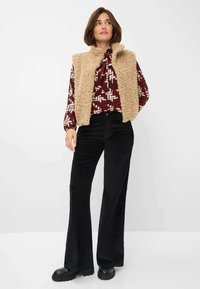 Beige nepbont vest over een geometric patroon in rood en crème blouse, gecombineerd met zwarte ribfluwelen uitlopende broek en chunky zwarte schoenen.