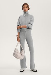 Siva flisasta zip-up majica z visokim ovratnikom, v kombinaciji z ujemajočimi se širokimi hlačami. Drži teksturirano krema torbico. Bele superge.