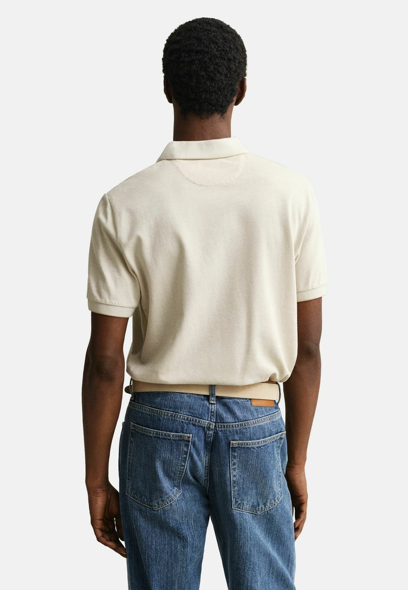 Man met kort krullend haar, gekleed in een beige polo met korte mouw, ingestopt in een blauwe spijkerbroek, staand met zijn rug naar de camera tegen een witte achtergrond.