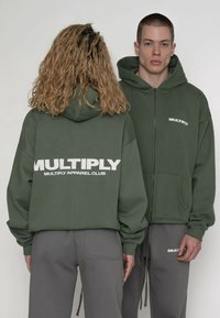 Olivengrøn hættetrøje med lynlås og stort hvidt "MULTIPLY"-logo på ryggen og fronten, med rummelig hætte og løs pasform. Kombineret med grå bukser.