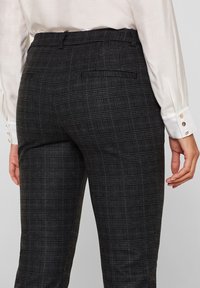 Vrouw in een donkergrijze geblokte broek met achterzakken en een witte blouse met lange mouwen en geknoopte manchetten, van achteren gezien.