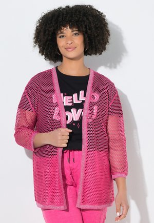 Vrouw met krullend haar die een zwart shirt met roze tekst draagt, een roze mesh vest en roze broek tegen een witte achtergrond.
