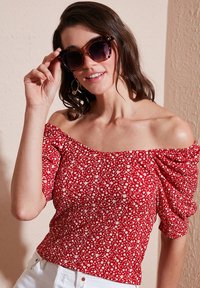 Red floral off-the-shoulder blouse with puffed sleeves, featuring a white flower pattern. Model wears tortoise-shell sunglasses and white pants.