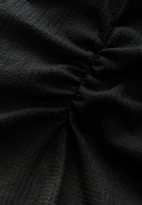 Schwarzer Stoff mit einer strukturierten, zerknitterten Oberfläche, die in der Mitte geraffte Details aufweist. Keine deutlichen Muster oder Akzente sichtbar.