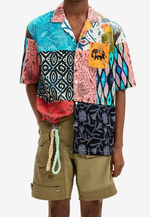 Homme portant une chemise à manches courtes patchwork multicolore avec une poche ornée d'un motif d'éléphant, un short cargo olive, et des porte-clés tressés suspendus à une passants de ceinture.