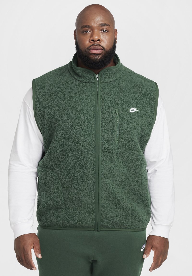 Colete de fleece verde com colarinho alto, fecho em zipper, bolsos laterais e um pequeno logótipo branco da Nike no peito. Usado sobre uma camisa de manga longa branca.