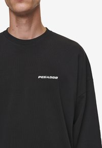 Fekete, hosszú ujjú póló, 100% pamutból készült, fehér hímzett "PEGADOR" logóval a bal mellen. Kerek nyakkivágású dizájn.